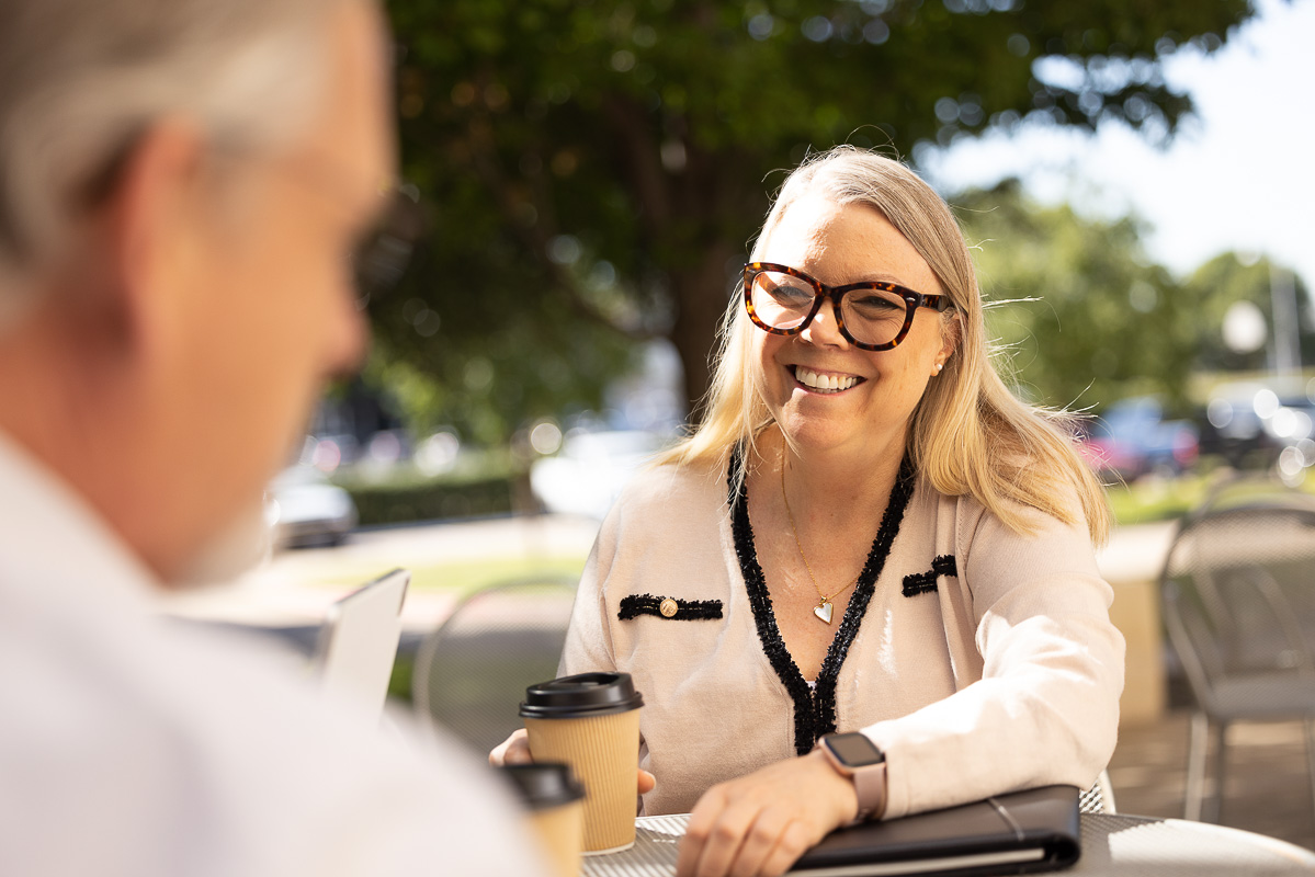 woman with glasses sitting at table outside smiling while holding coffee cup