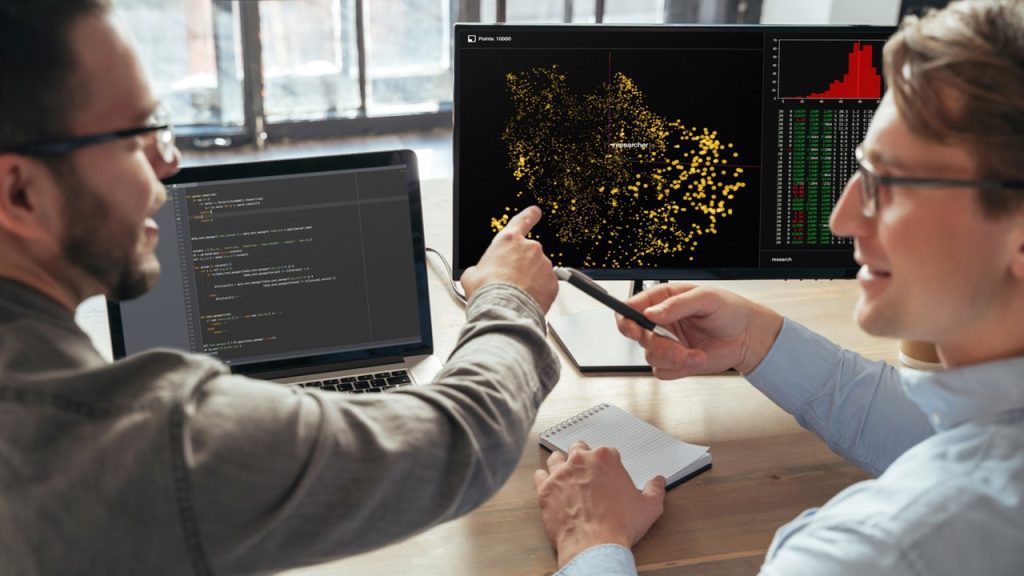 Closeup of two machine learning specialists descanting about new project in office in front of pc, analyzing data. Selective focus on computer screen